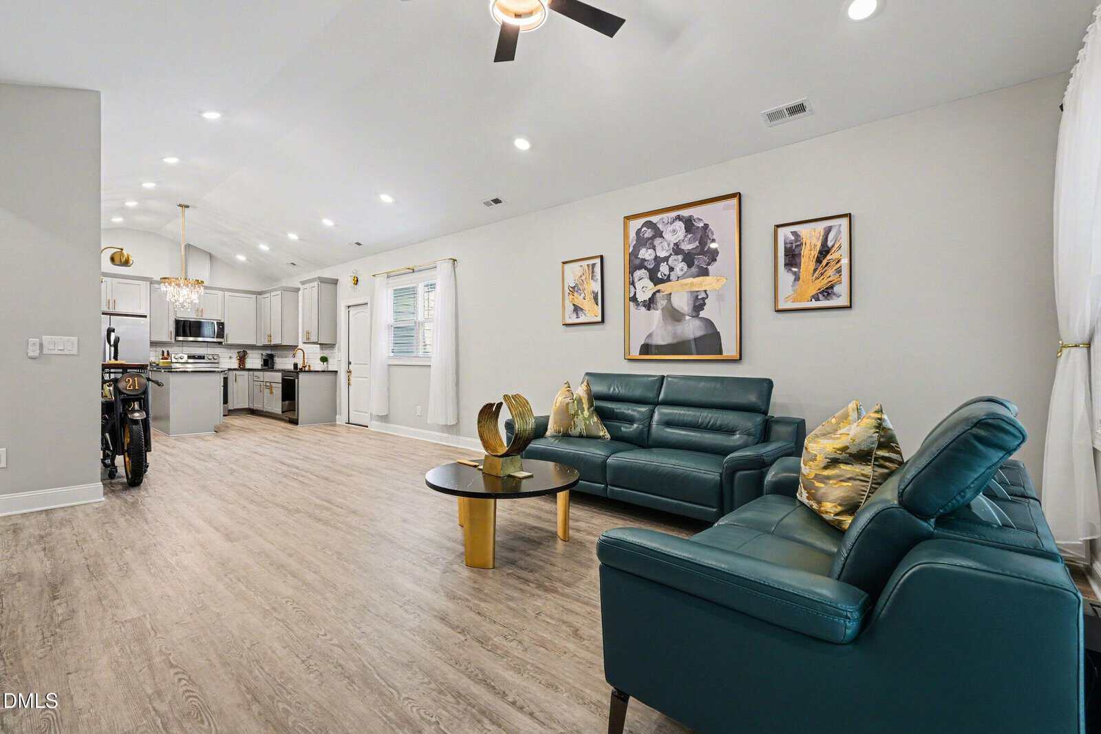 Modern living room with teal sofas, a round glass coffee table, wood flooring, and abstract art on the walls. The space opens into a bright kitchen with white cabinets and stainless steel appliances.
