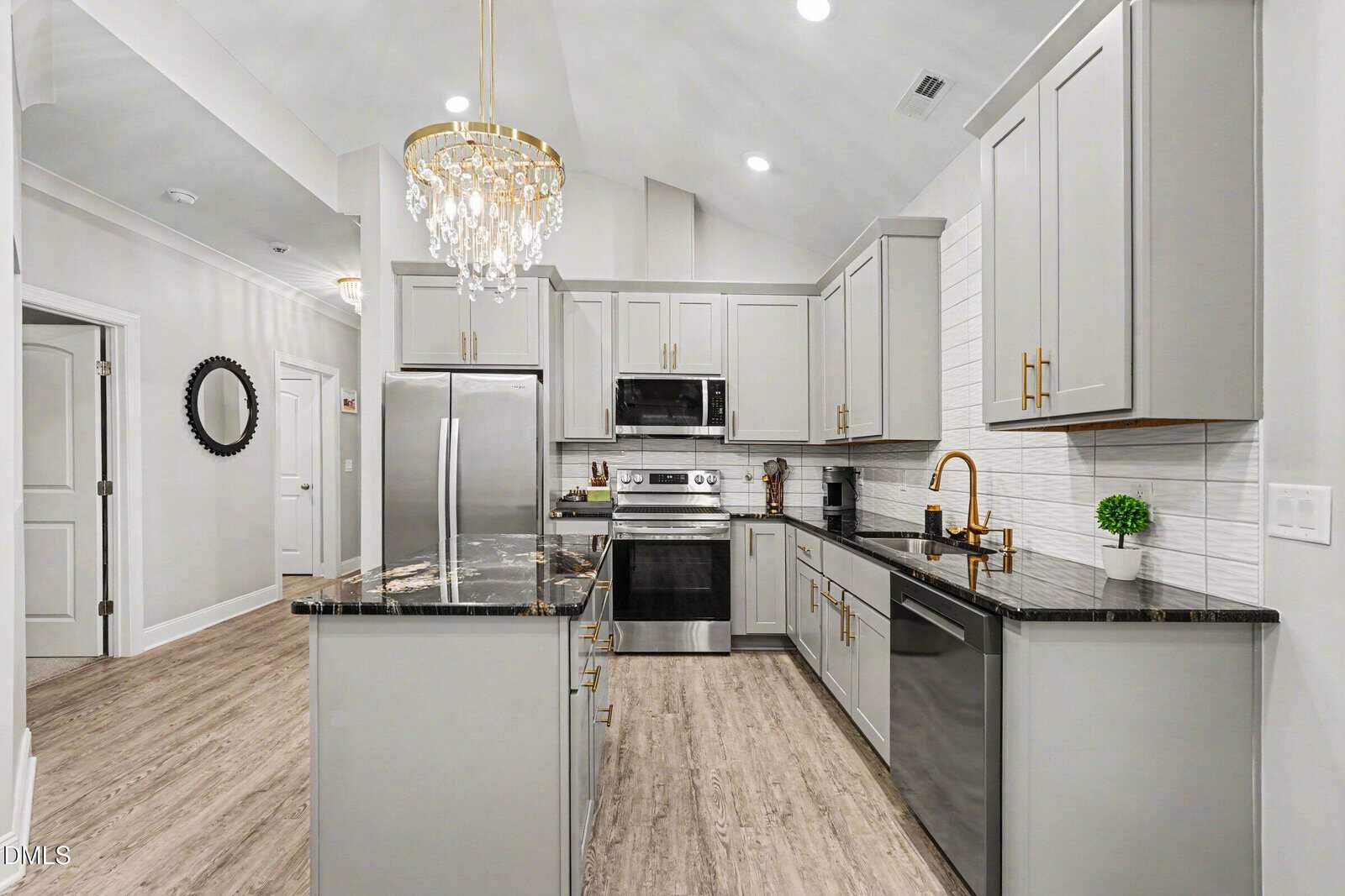 Modern kitchen with gray cabinets, black countertops, stainless steel appliances, gold fixtures, a chandelier, light wood flooring, and white tile backsplash. Small green plant on the counter near the sink.