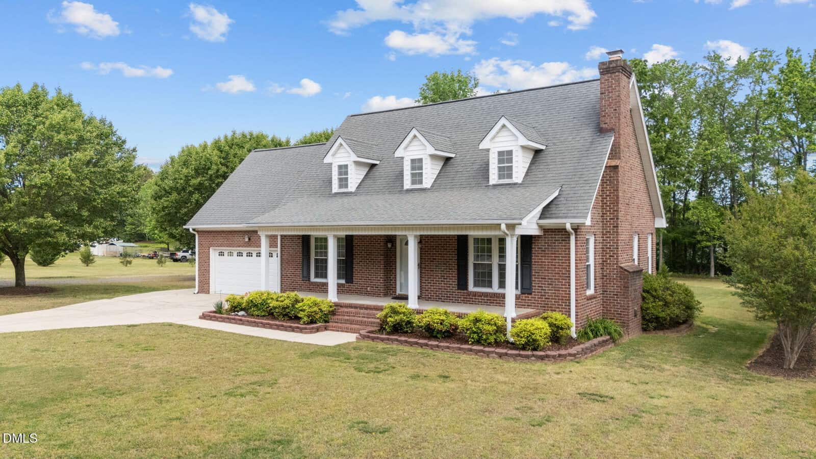 A brick two-story house with a gray roof and three dormer windows, a front porch with white columns, a double garage, and neatly trimmed shrubs, set on a spacious green lawn with trees in the background under a blue sky.