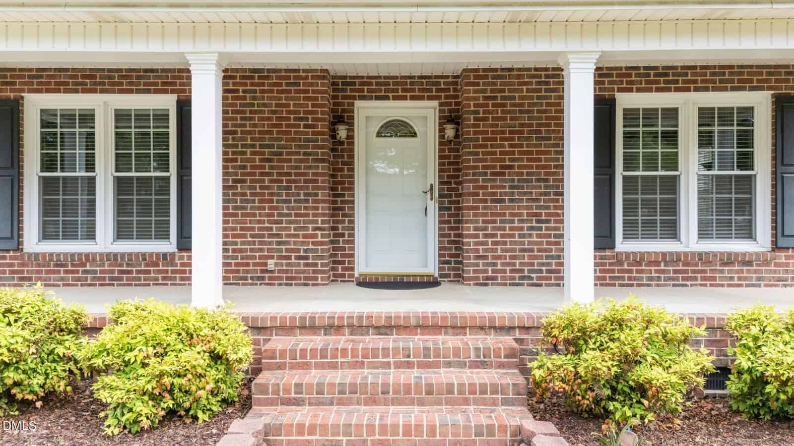 A white front door with a semicircular window sits between two columns on a red brick house. There are two windows with black shutters on either side and a set of brick steps with bushes in front.