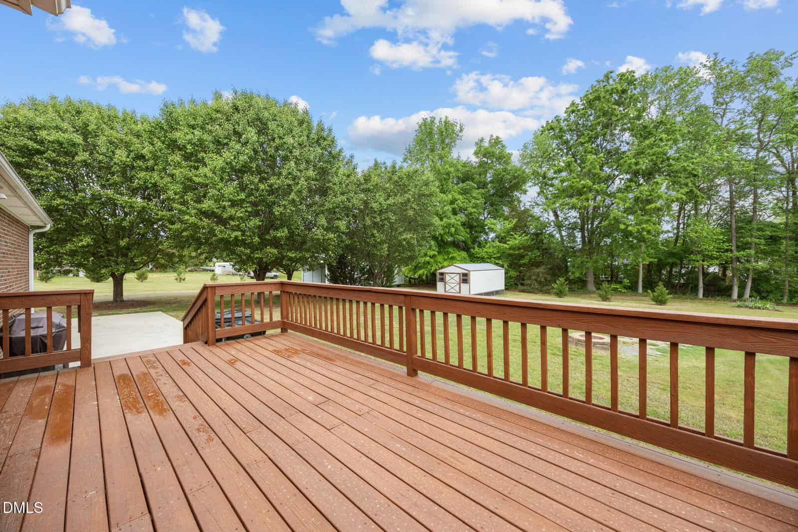 A spacious wooden deck with railings overlooks a large grassy backyard with mature trees and a white storage shed under a partly cloudy sky.