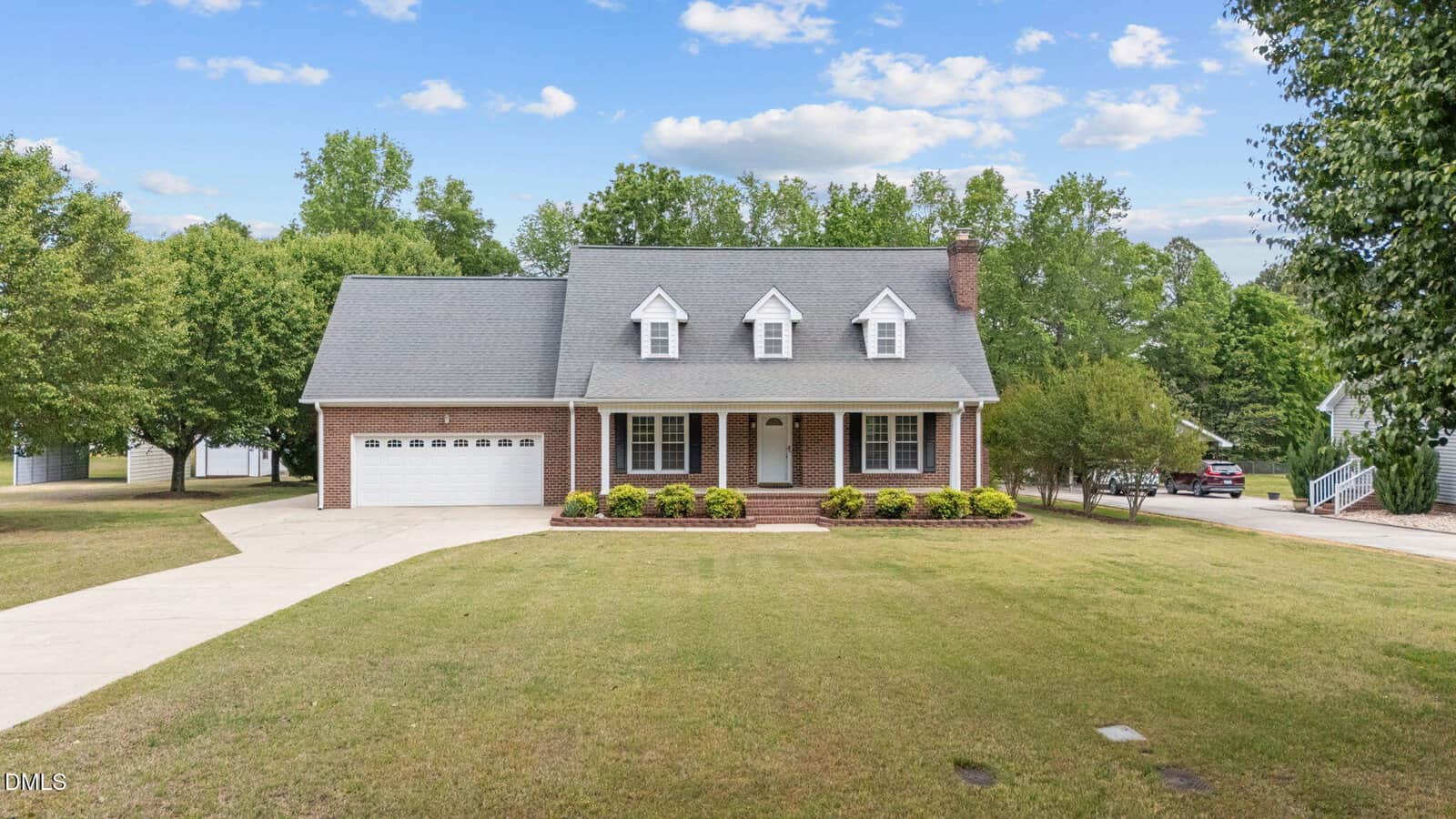 A two-story brick house with dormer windows, an attached two-car garage, a front porch, and a well-maintained lawn, surrounded by trees under a partly cloudy sky.