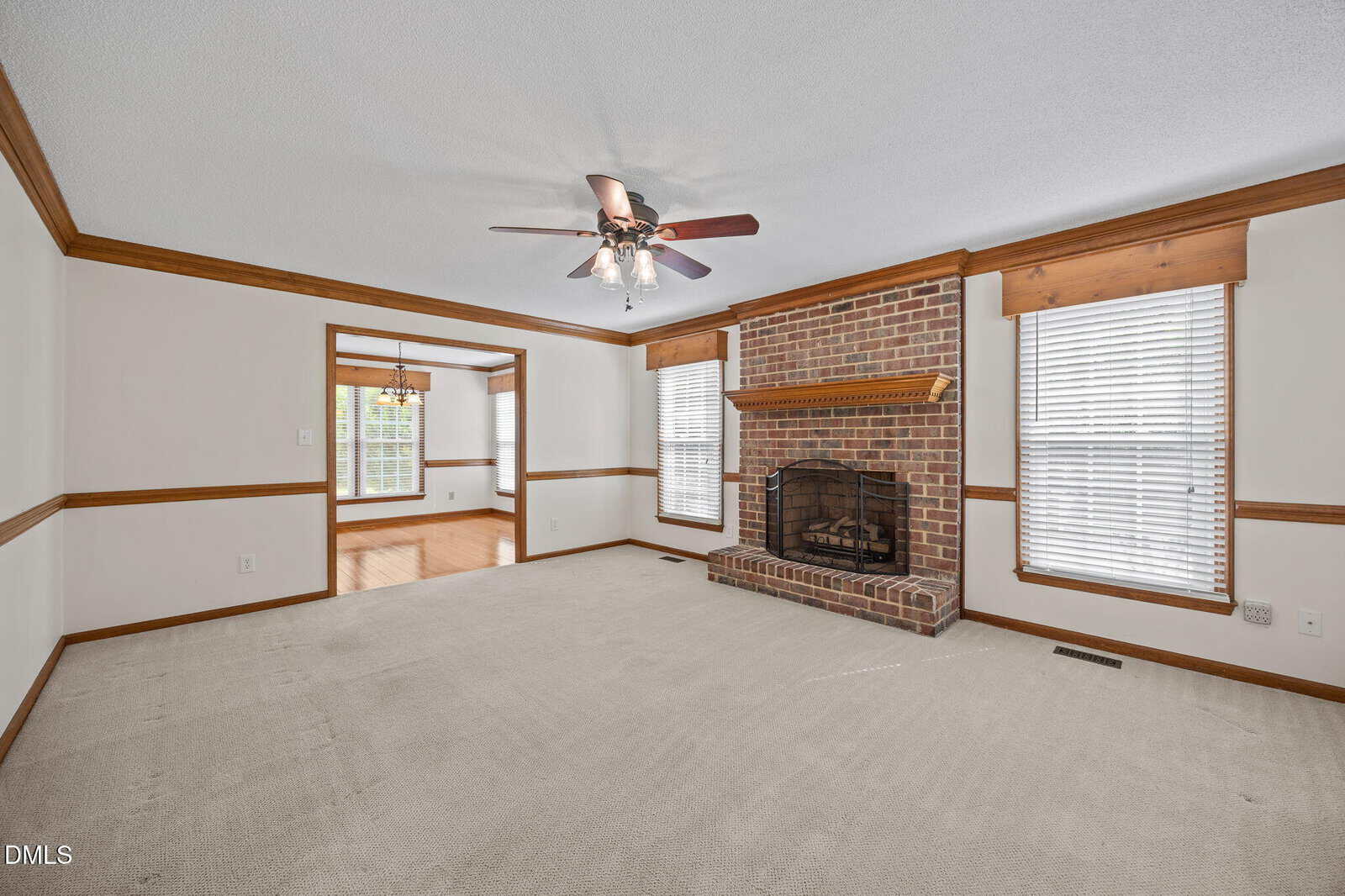Spacious living room with beige carpet, white walls, wood trim, brick fireplace with wooden mantle, ceiling fan with light, and large windows with wood blinds. Adjoining dining area visible through wide doorway.