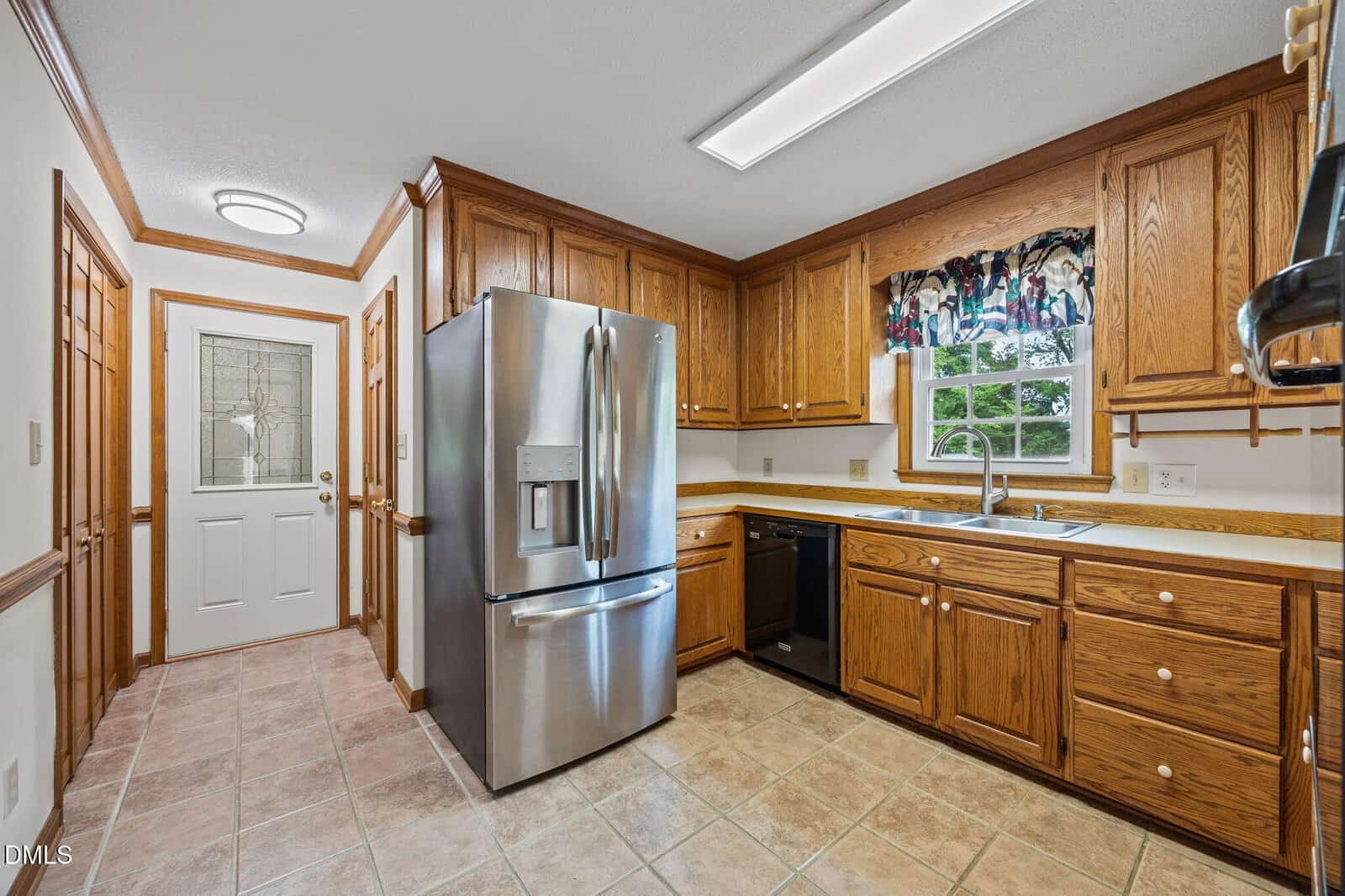 A kitchen with wooden cabinets, stainless steel refrigerator, tiled floor, a window above the sink, and a white door with a glass panel leading outside.