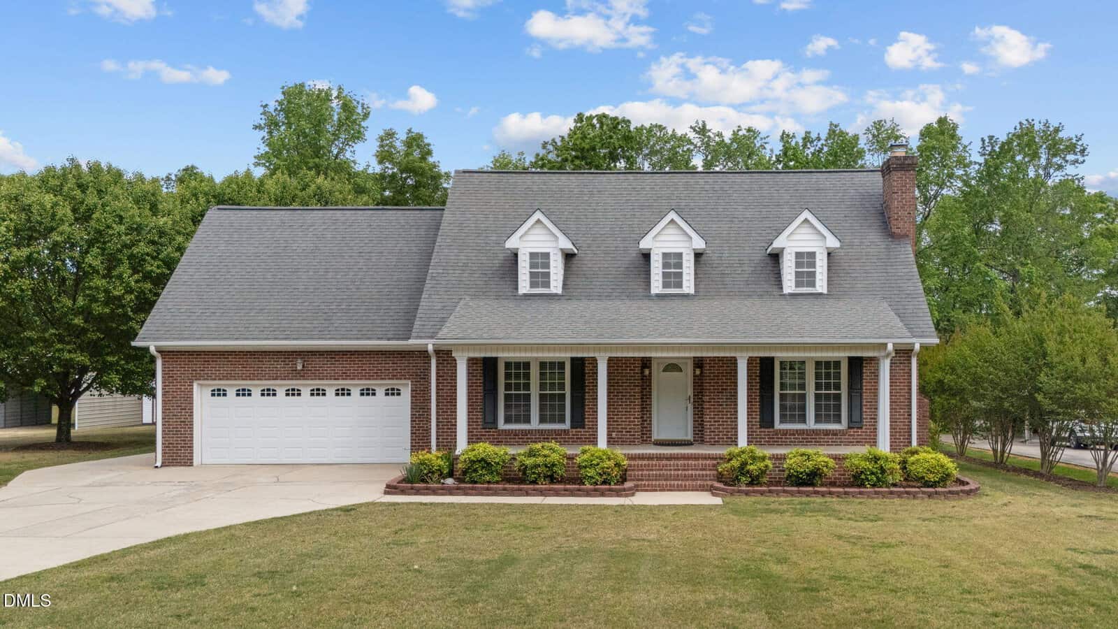 A brick house with gray roof, white trim, three dormer windows, a front porch, two-car garage, small shrubs, and a spacious front yard under a blue sky with scattered clouds.
