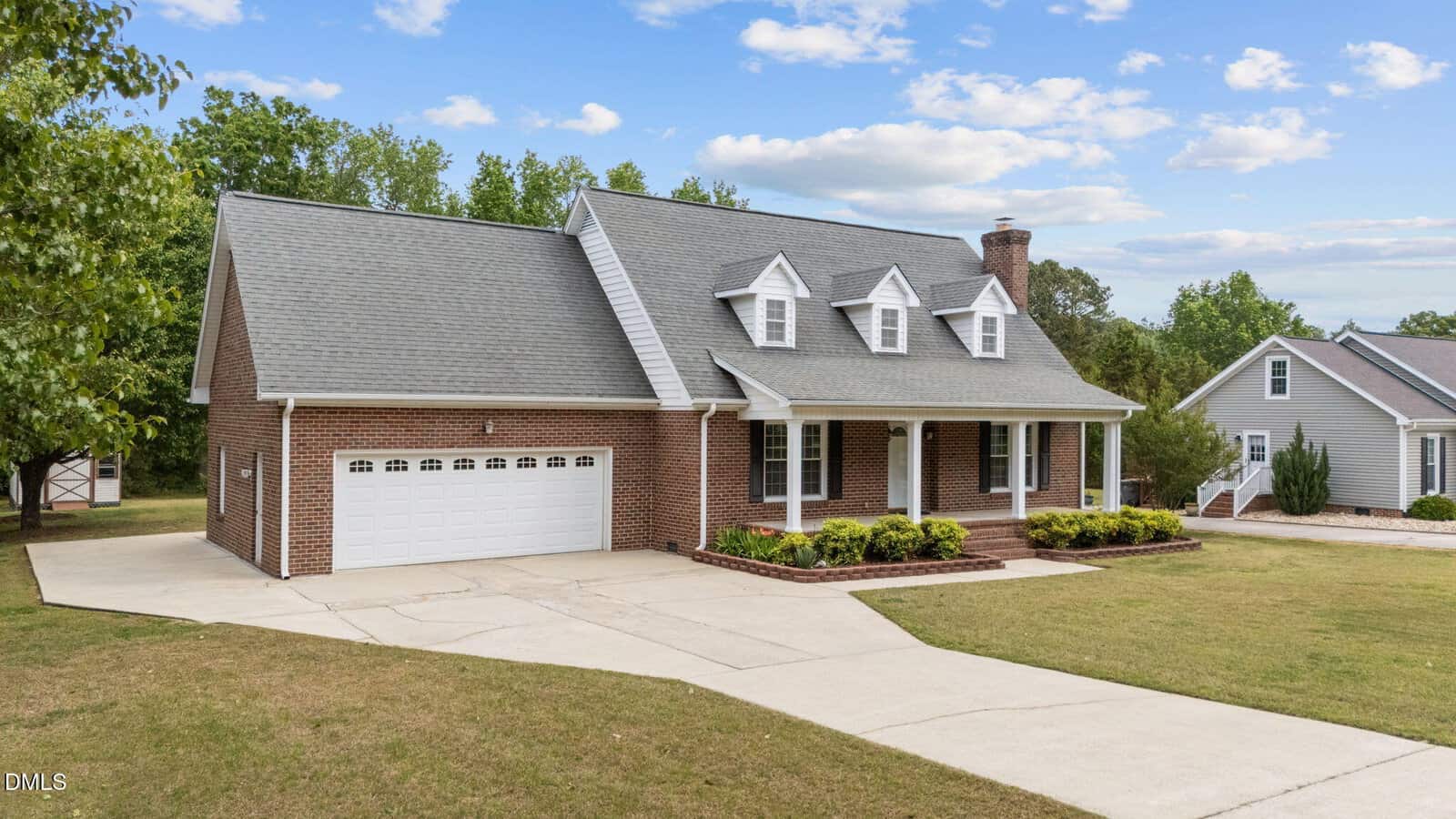 A red brick house with a gray roof, white trim, three dormer windows, a front porch, and a two-car garage. The home sits on a spacious lawn with a curved driveway and trees in the background under a blue sky.