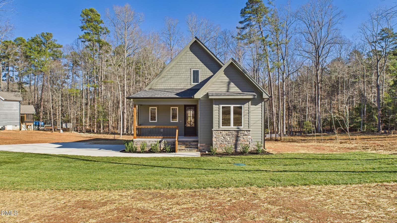 A small, modern gray house with a front porch, gabled roof, and stone accents sits on a spacious lot with a fresh green lawn, concrete driveway, and surrounded by tall trees under a clear blue sky.