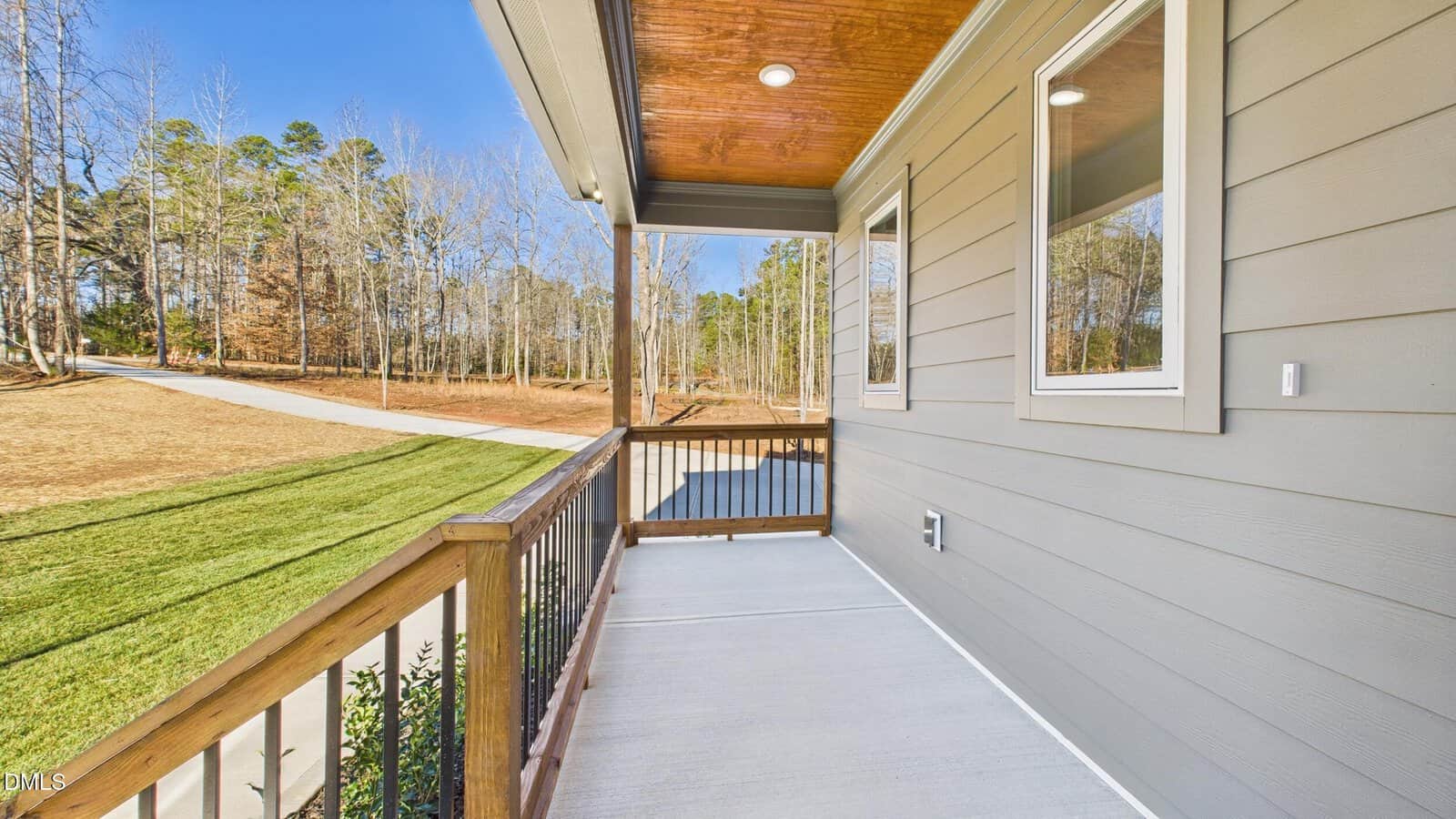 A small, covered front porch with wooden railings and gray siding overlooks a driveway, green lawn, and trees on a sunny day.