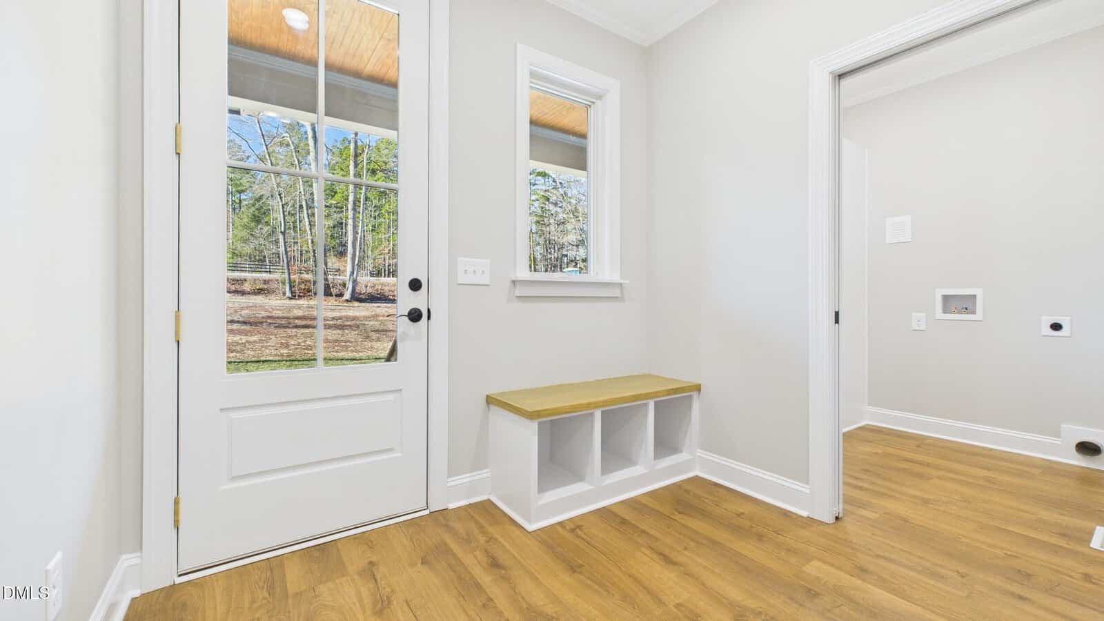 A bright mudroom with wood flooring, a white door with glass panes, a window, a small built-in bench with storage, and a laundry area visible through an open doorway.