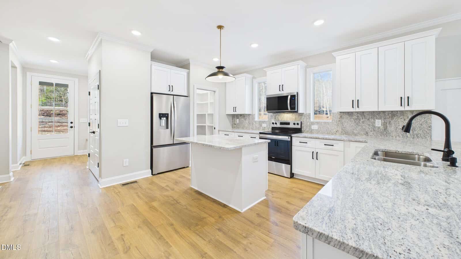 Bright modern kitchen with white cabinets, stainless steel appliances, a central island, granite countertops, wood flooring, and large windows letting in natural light. Door leads outside, and a black matte faucet sits above a sink.