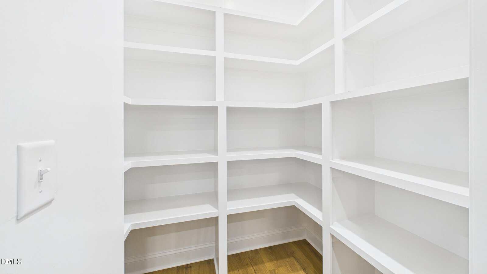 White walk-in pantry with built-in shelves on three walls, empty and well-lit. The floor is wooden, and a light switch is visible on the left wall.