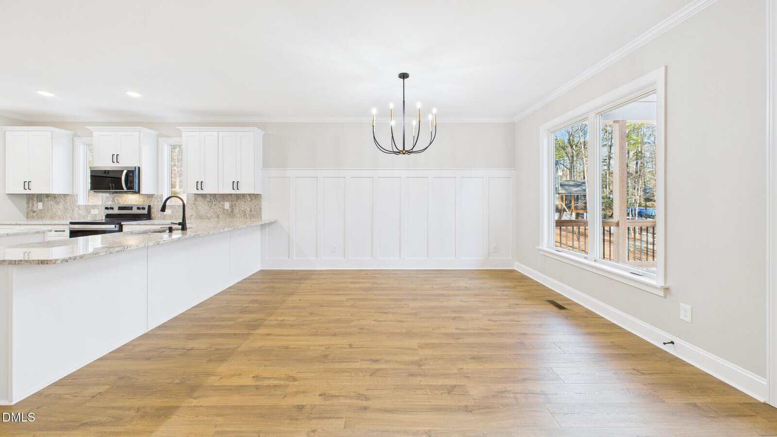 Bright, modern kitchen and dining area with white cabinets, granite countertops, wood floors, a large window, and a black chandelier. The space is open and filled with natural light.
