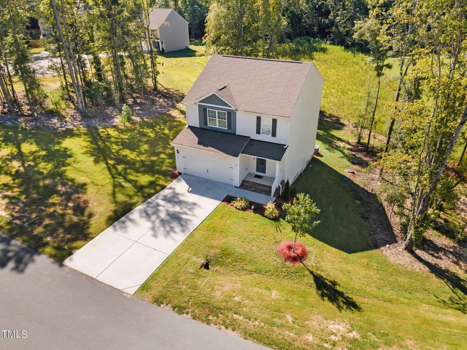 A two-story white house with a gray roof, attached garage, and neatly landscaped yard sits on a sunny corner lot surrounded by trees and green grass, viewed from above.