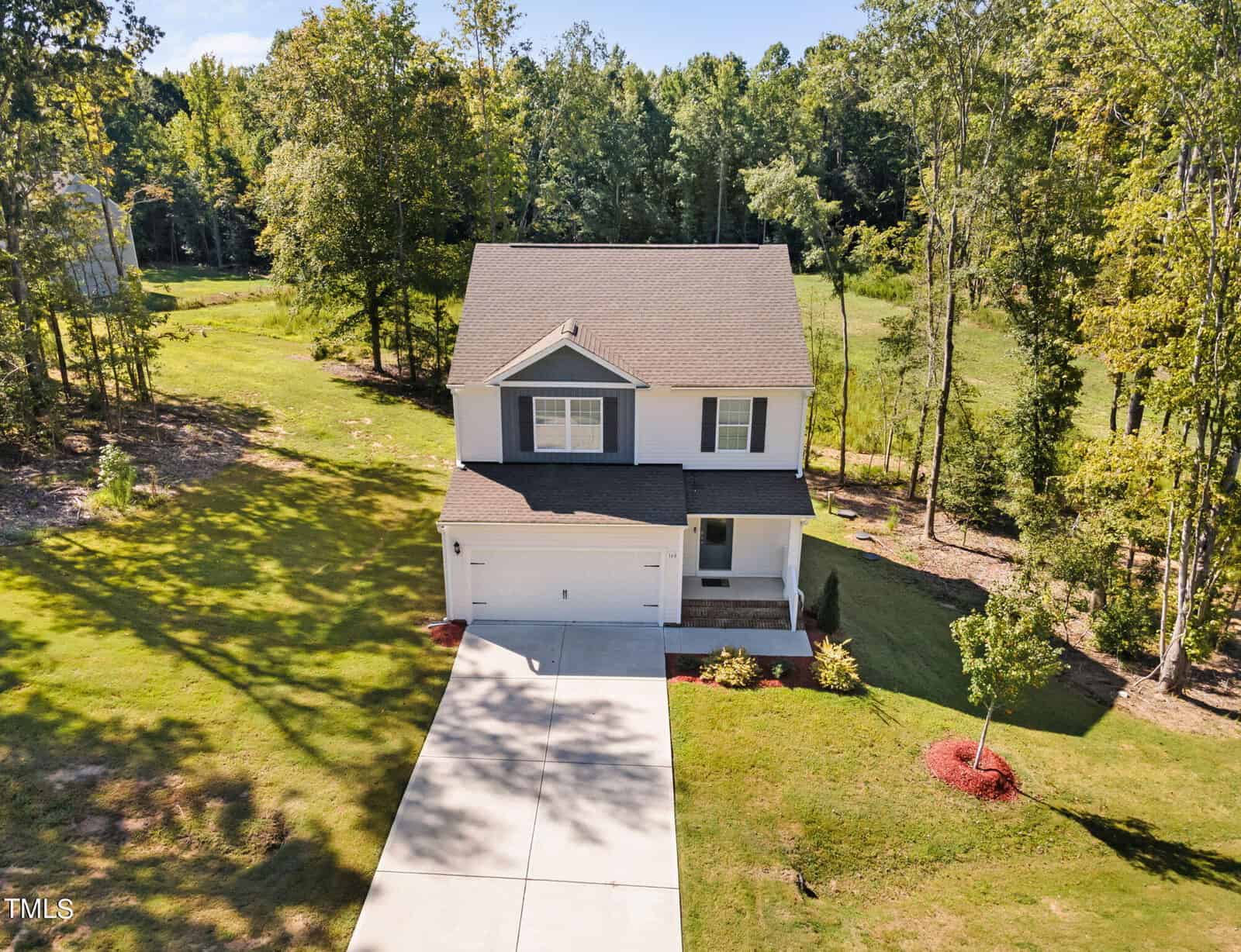 A two-story white house with a front porch and attached garage sits on a spacious, grassy lot surrounded by trees. A long driveway leads to the garage, and landscaping with mulch accents the front yard.