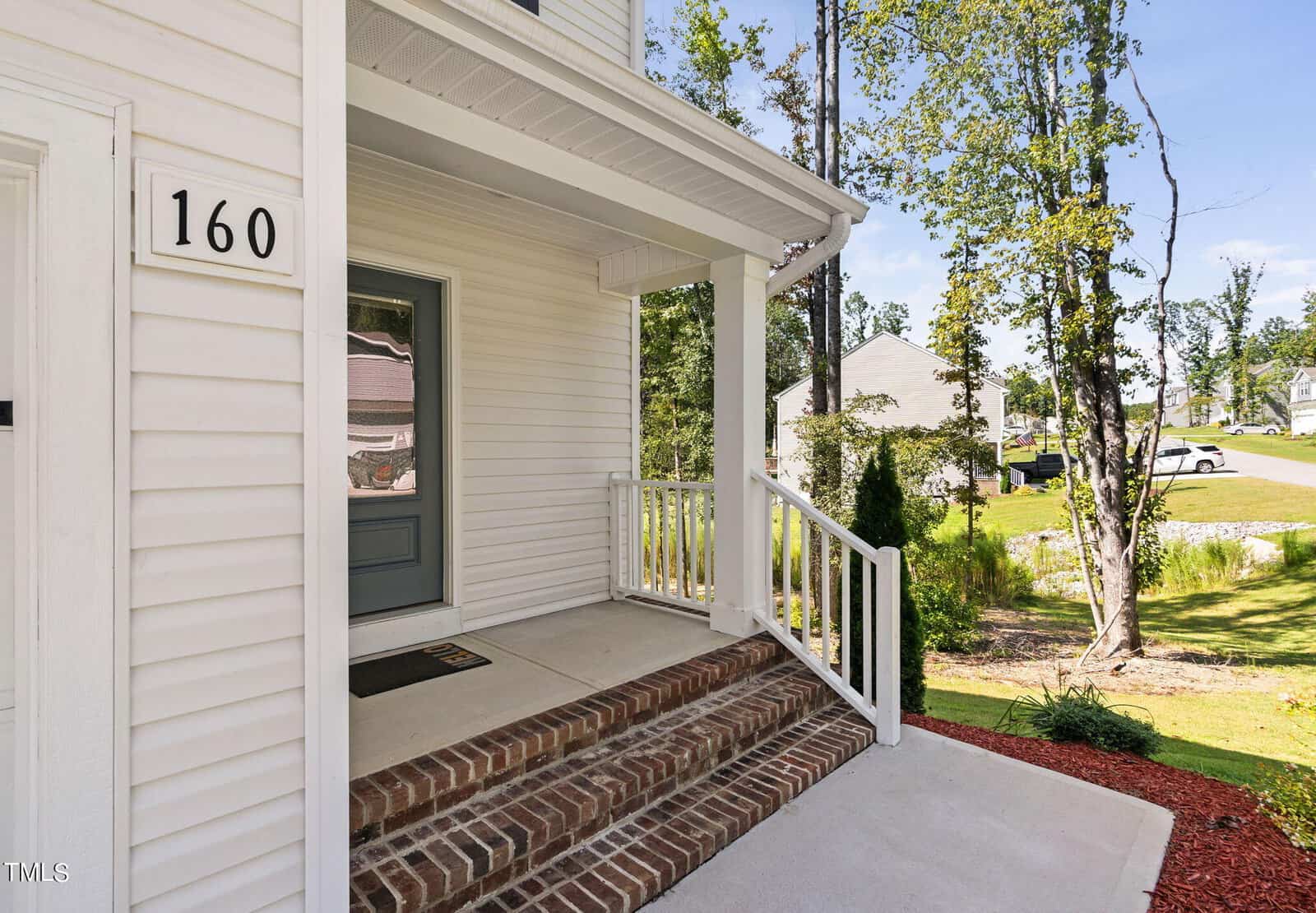 A front porch of a white house with a gray door, brick steps, white railings, and a house number 160. The yard has mulch and trees, with a suburban neighborhood visible in the background.