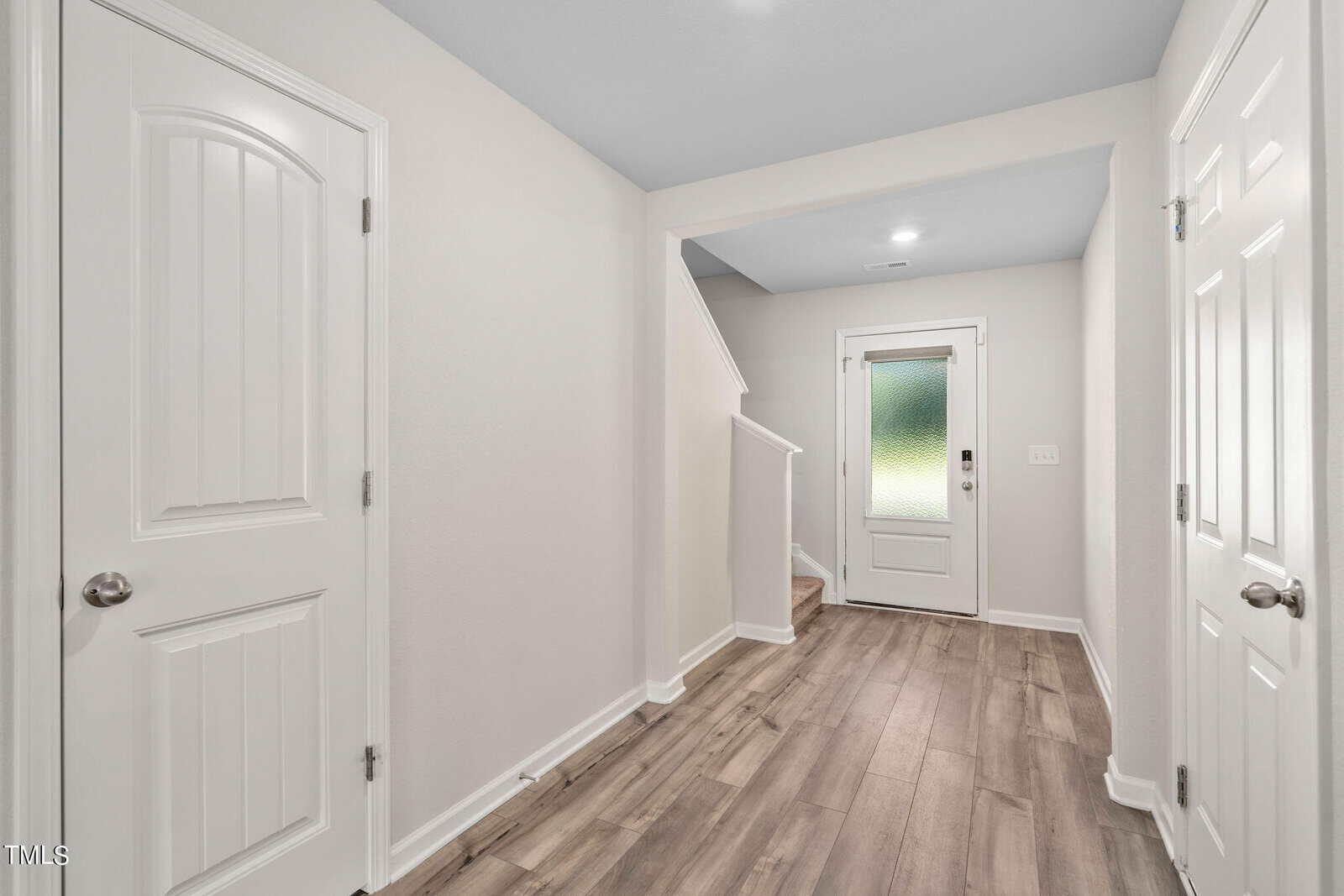 A bright, modern hallway with light-colored walls, wood-look flooring, two closed white doors on the left and right, a staircase leading up, and a back door with frosted glass at the end.