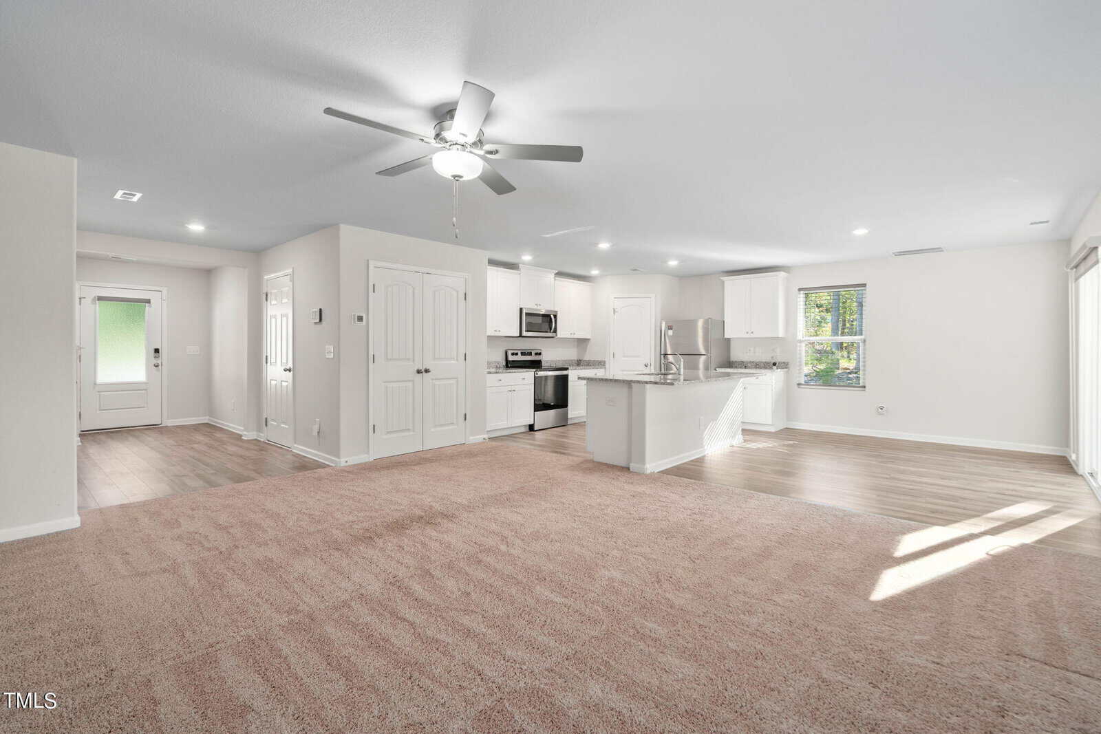 Open-concept living area with beige carpet, a ceiling fan, and adjacent kitchen featuring white cabinets, stainless steel appliances, and a breakfast bar. Sunlight streams through windows and a glass door.