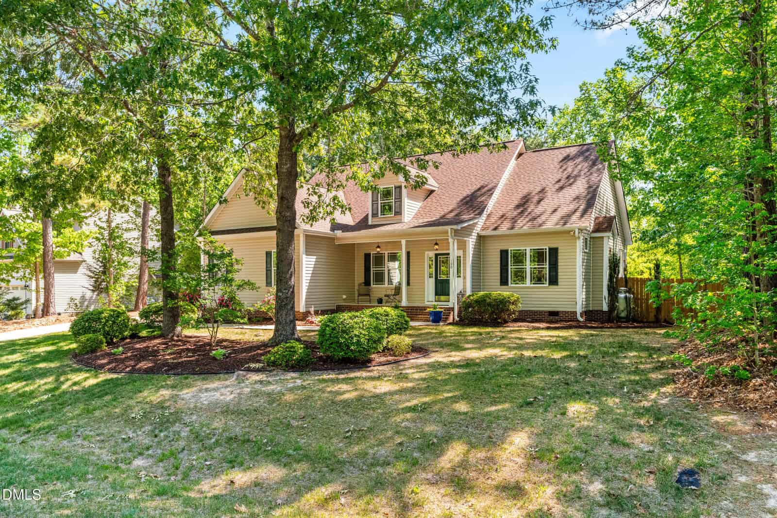 A light-colored house with a brown roof sits among tall green trees and trimmed bushes. The front yard has patchy grass, a mulched garden bed, and a covered porch with white columns. Sunlight filters through the leaves.