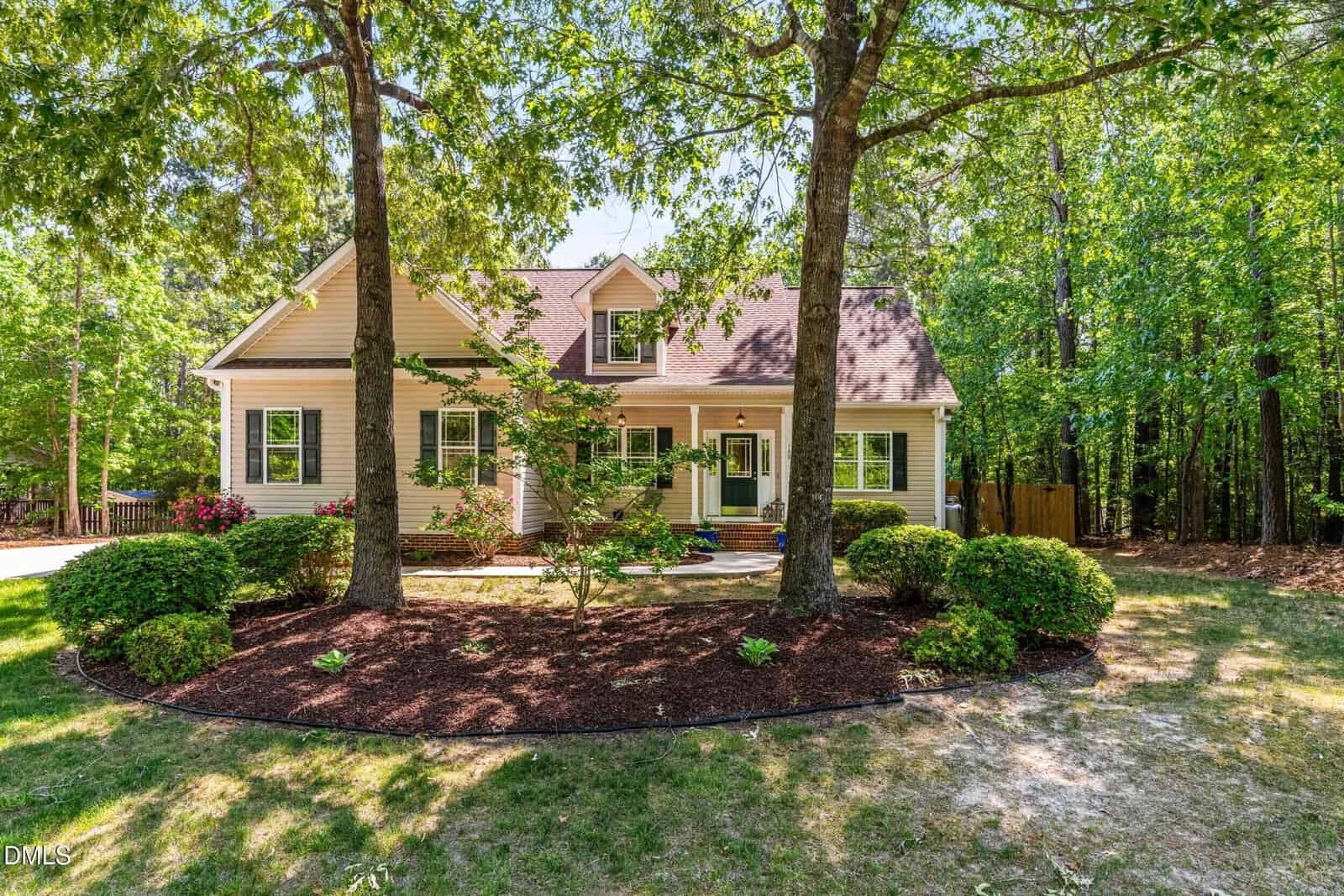 A beige two-story house with a front porch, surrounded by trees and neatly landscaped bushes under a clear sky. Sunlight filters through the foliage, highlighting the home's inviting appearance.
