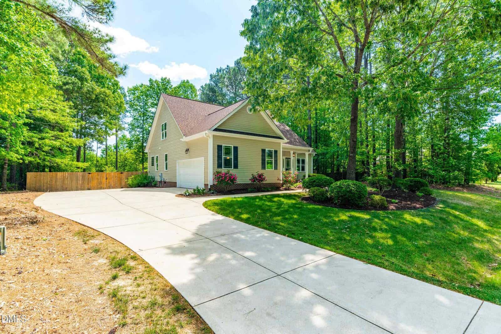 A light-colored house with a gabled roof and front porch sits by a curved driveway, surrounded by green trees, shrubs, and a well-manicured lawn on a bright sunny day.