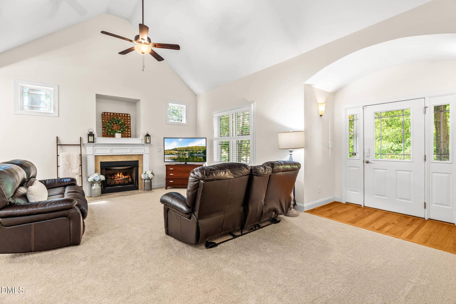 Bright living room with high ceiling, brown leather sofas, a fireplace, TV, and large windows near a white double front door. Neutral décor with carpeted floor and wooden accents.