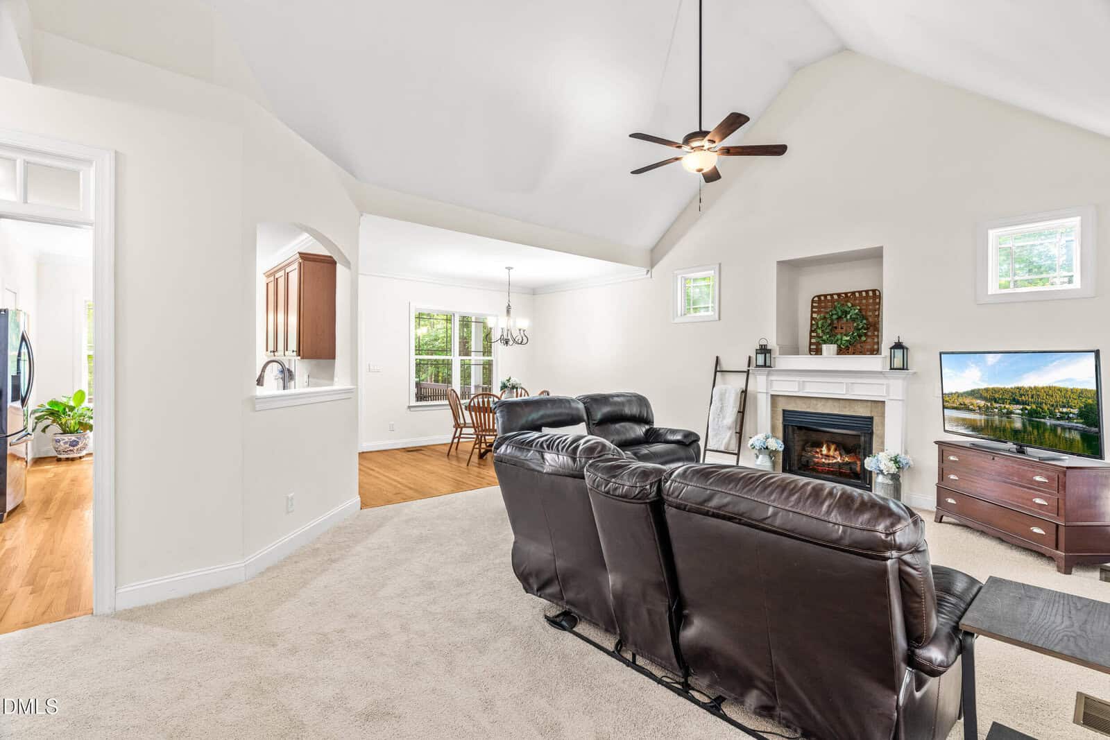 Bright living room with vaulted ceiling, ceiling fan, fireplace, wall-mounted TV, dark leather sofas, beige carpet, and an open view of the dining area and kitchen with hardwood floors.