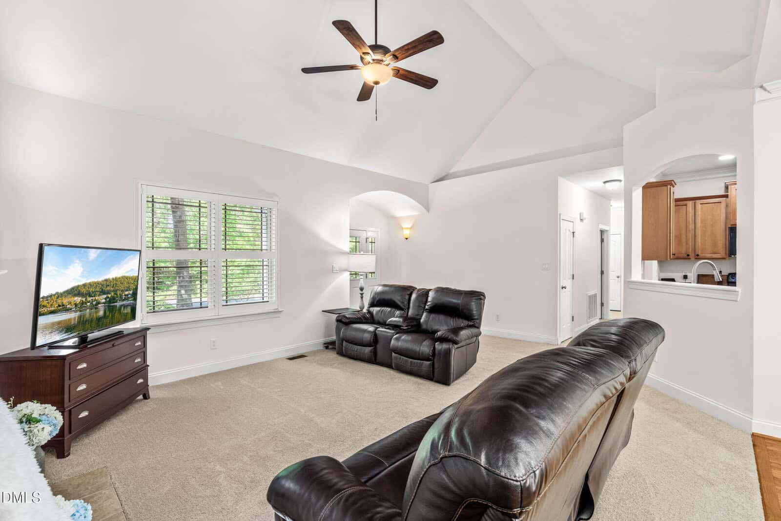 A bright living room with beige carpet, two dark leather sofas, a ceiling fan, a TV on a wooden stand, and windows with white shutters. The kitchen is visible through an open archway.