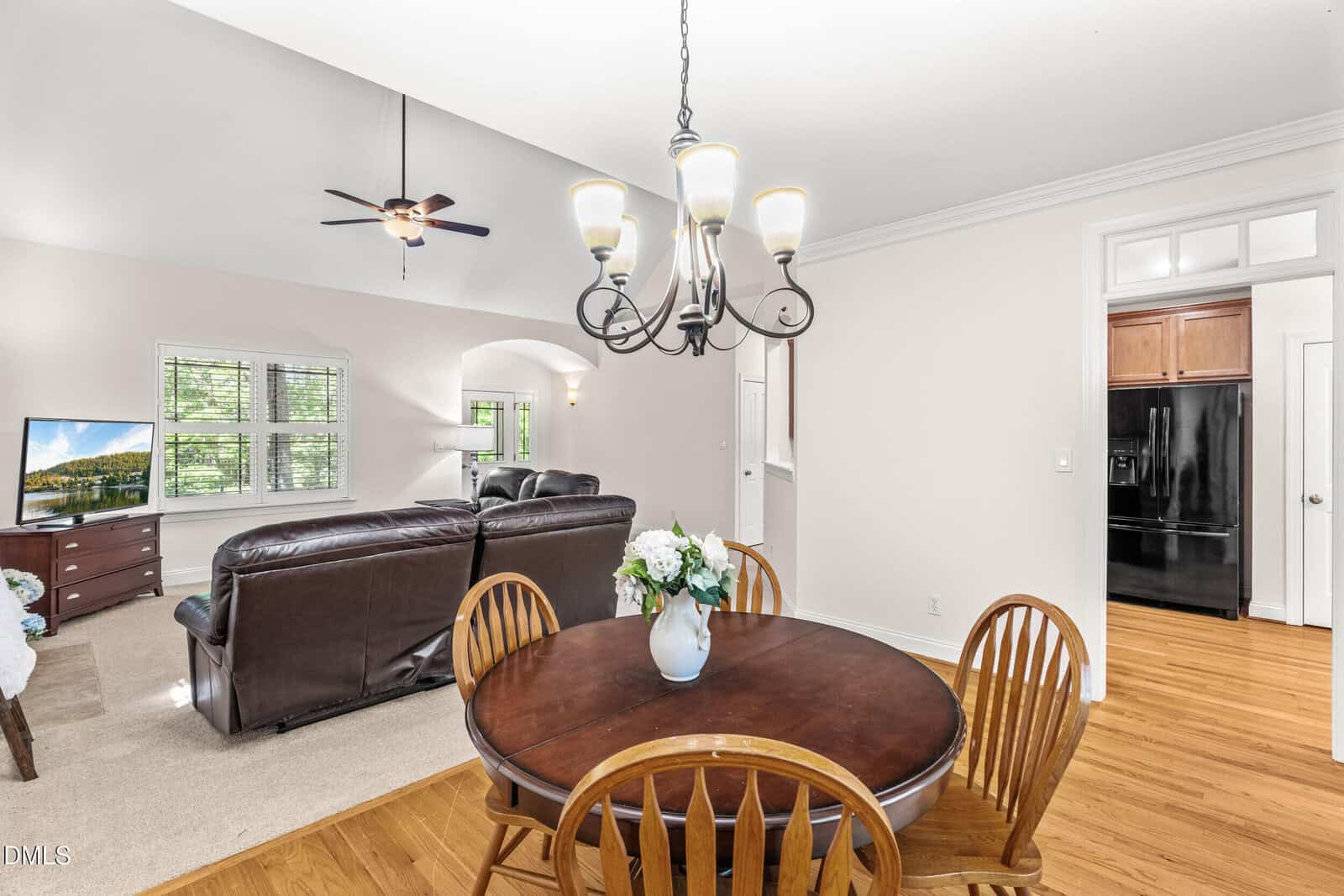 Bright dining area with a round wooden table and four chairs, adjacent to a living room with leather sofas, TV, and ceiling fan. Kitchen with dark appliances visible through an open doorway. Hardwood floors and white walls.