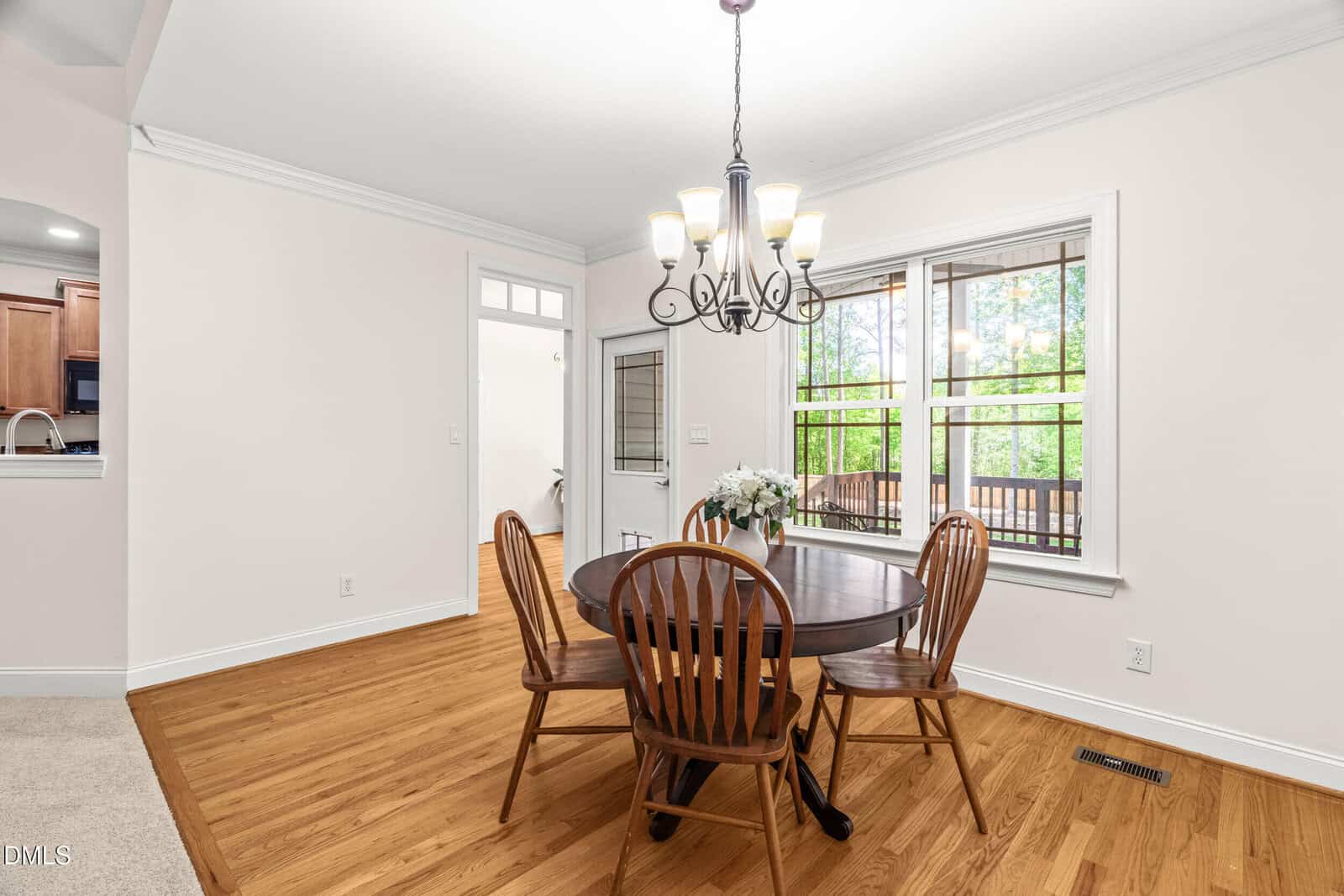 A bright dining area with a round wooden table, four chairs, a floral centerpiece, hardwood floors, a chandelier, large windows, and a view into a kitchen and another room.