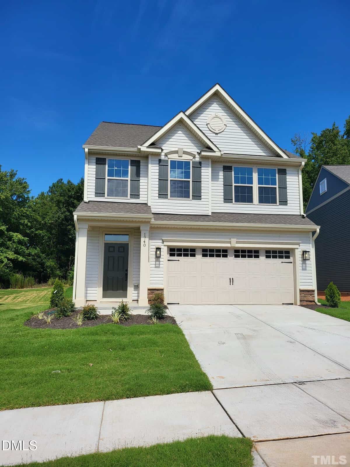 Two-story suburban house with light-colored siding, dark shutters, and a double garage. Manicured lawn and young shrubs in front, with a concrete driveway and blue sky overhead.
