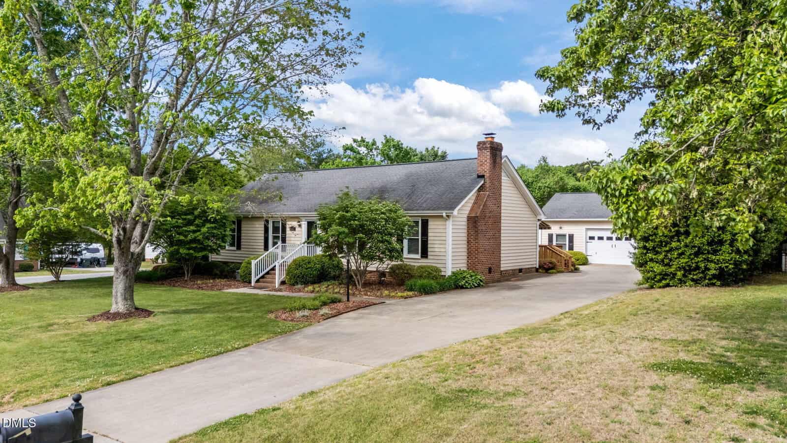 A single-story house with light siding, a brick chimney, and a large front porch sits amid green trees and a manicured lawn. A long driveway leads to a detached garage in the background.