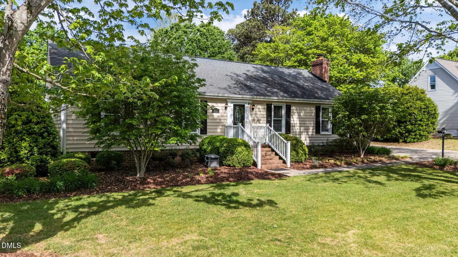 A single-story beige house with white trim, black shutters, and a brick chimney sits surrounded by green trees and bushes. Steps lead to the front door, and the lawn is neatly landscaped under a partly cloudy sky.