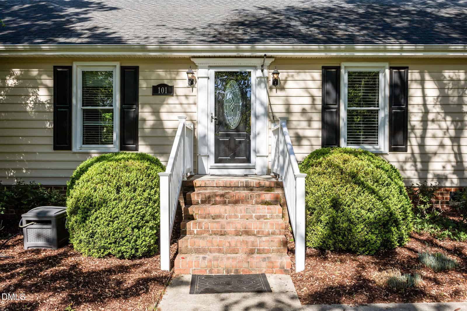 Front view of a house with beige siding, a black front door with glass panel, brick steps, white railings, black shutters, two windows, and neatly trimmed green bushes on either side of the entrance.