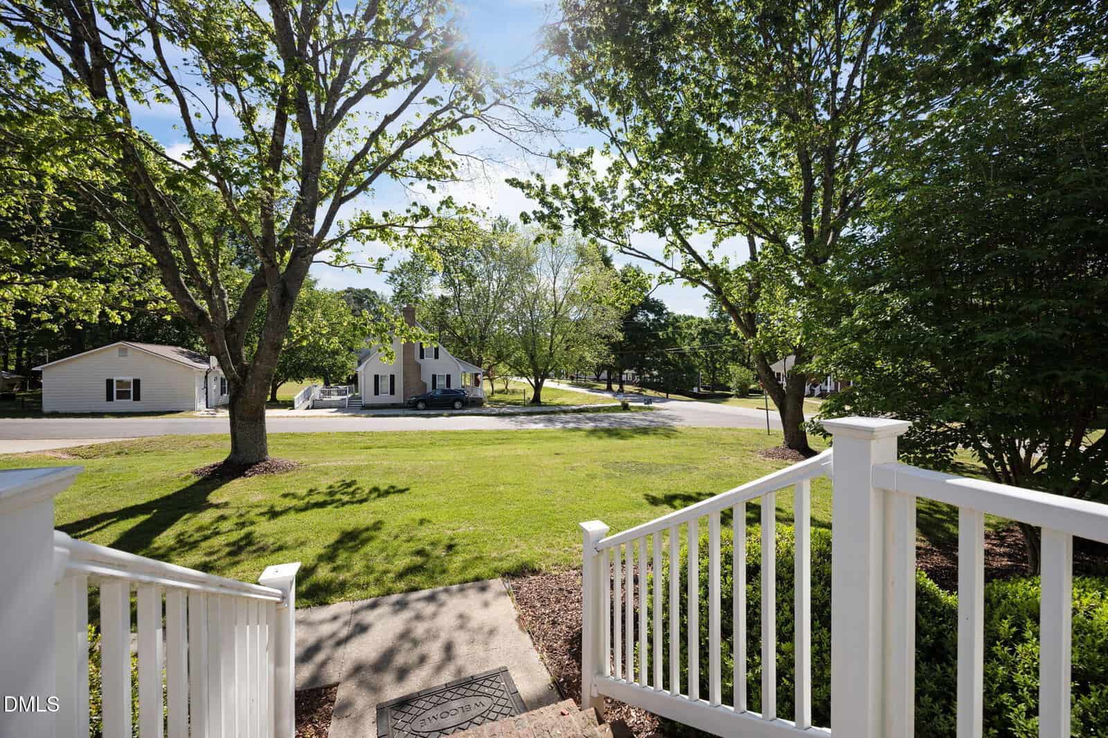 View from a porch with white railings, looking out onto a green lawn, sidewalk, and quiet suburban street lined with trees and white houses under a sunny sky.