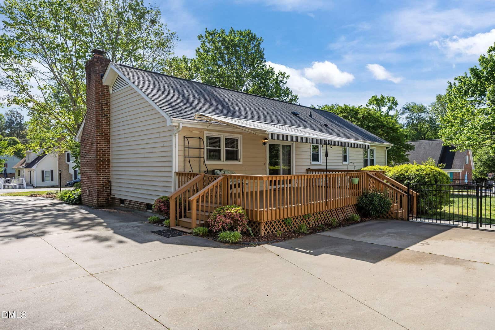 A single-story beige house with a dark roof and large brick chimney features a wooden ramp leading to the front door, surrounded by green shrubs and trees, on a sunny day with a clear blue sky.