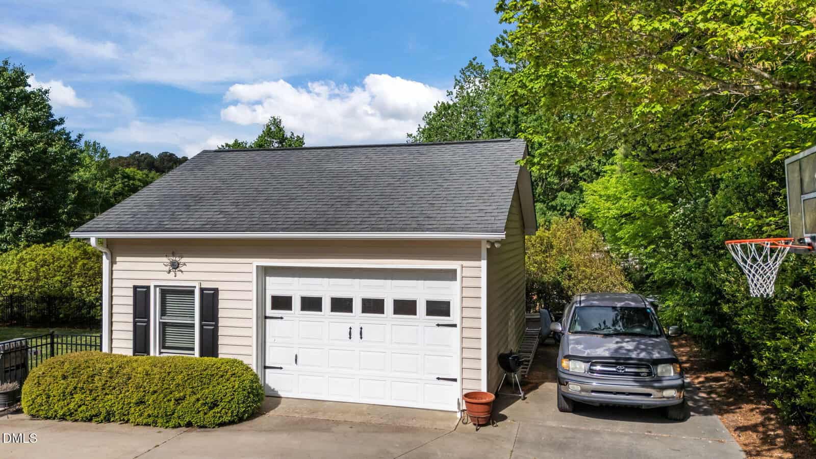 A beige detached garage with a white door, a black truck parked beside it, green bushes in front, and a basketball hoop on the right, surrounded by trees under a partly cloudy sky.