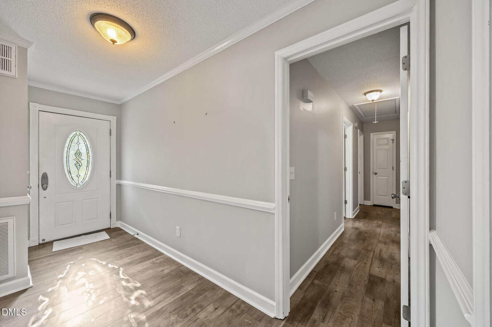 A bright hallway with wood flooring, light gray walls, white trim, and an oval glass front door. Two ceiling lights and multiple doors lead to other rooms. Sunlight shines through the front door onto the floor.