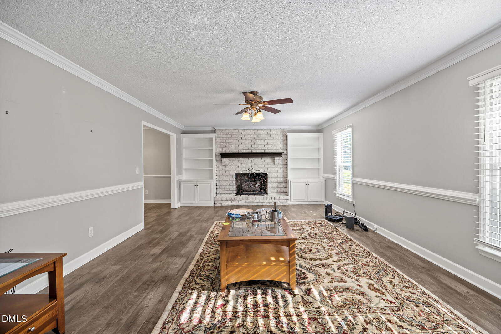 A living room with gray walls, white trim, wood floors, a patterned rug, a ceiling fan, a brick fireplace with shelves, a wooden coffee table, and windows letting in natural light.