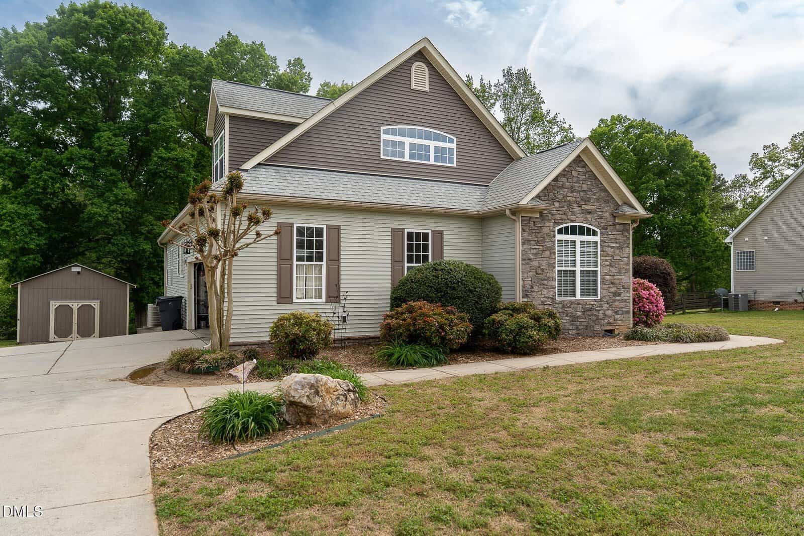 A modern two-story house with gray siding and stone accents, surrounded by shrubs and a well-kept lawn, with a small shed visible in the backyard and trees in the background.