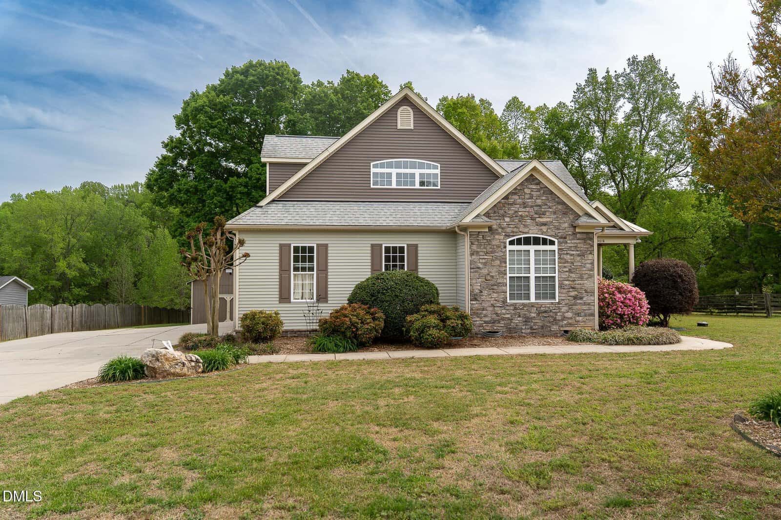 A two-story suburban house with stone and tan siding, surrounded by green trees, bushes, and a manicured lawn, under a partly cloudy sky. A curved driveway leads to the side of the home.