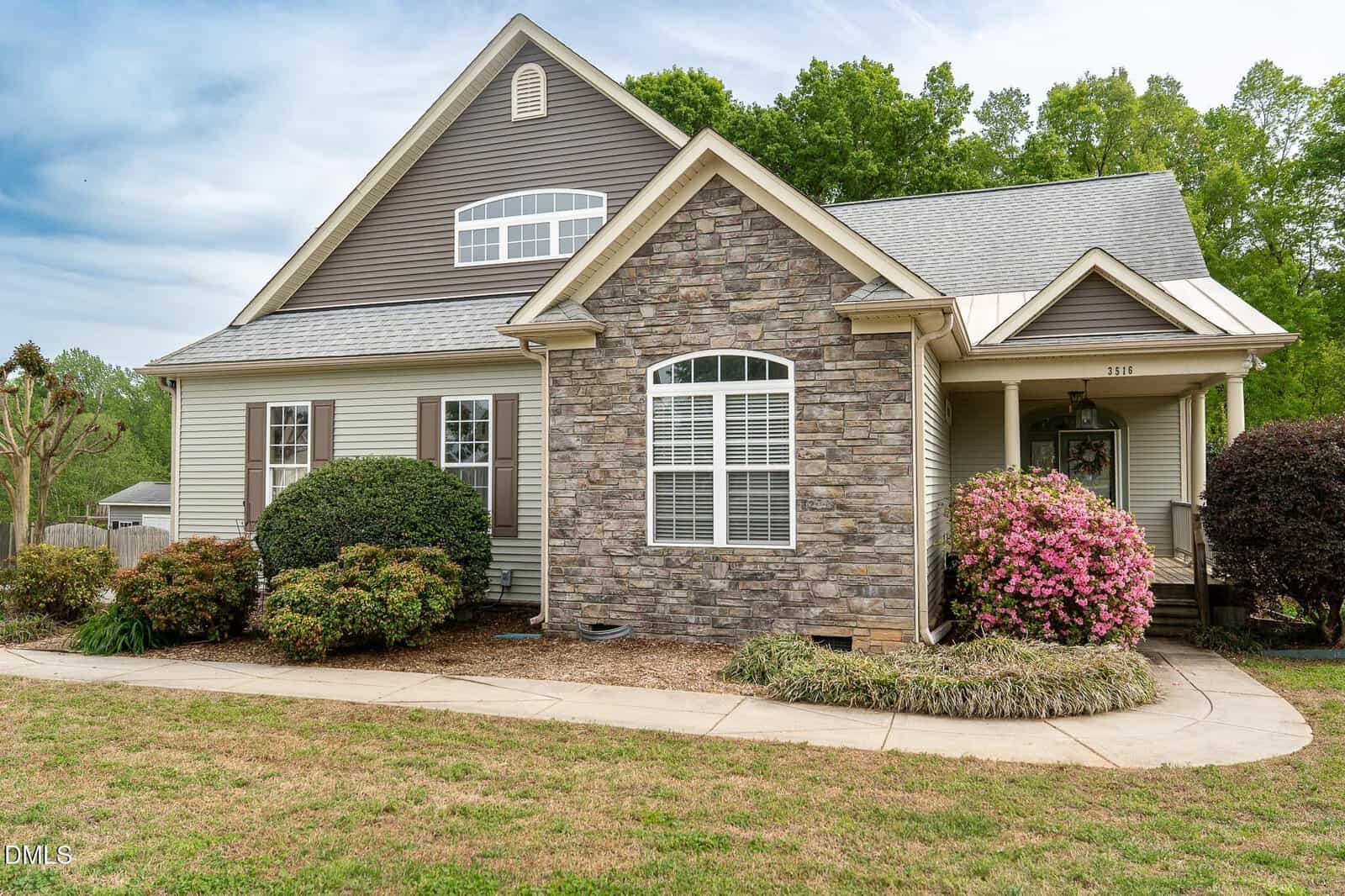 A suburban house with stone and siding exterior, gabled roof, white trim, well-kept shrubs, blooming pink flowers, and a grass lawn, set against a backdrop of trees and a blue sky.