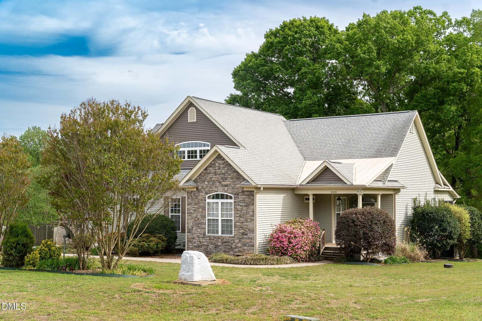 A two-story suburban house with stone and beige siding, surrounded by trees, bushes, and flowering plants under a partly cloudy sky.