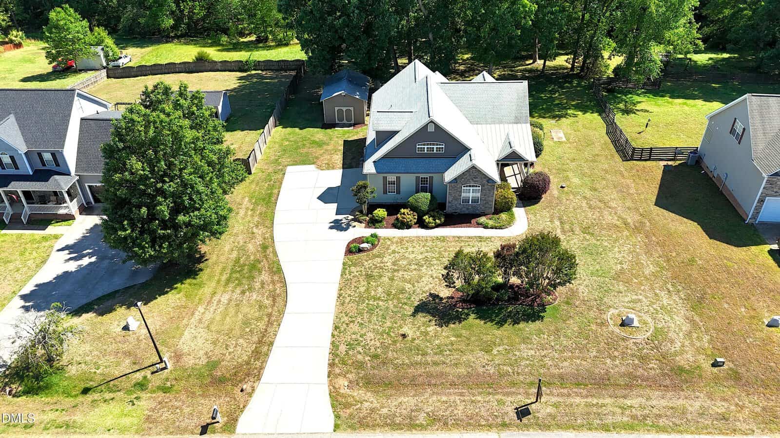 Aerial view of a gray two-story house with a large front yard, curved driveway, detached shed in the backyard, and surrounding trees and neighboring homes.