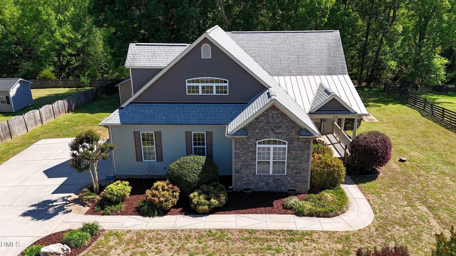 Aerial view of a two-story house at 3516 Bluebonnet Drive, Wake Forest, with gray siding and stone accents, landscaped shrubs and trees, a curved driveway, a fenced backyard, and lush green trees surrounding the property.
