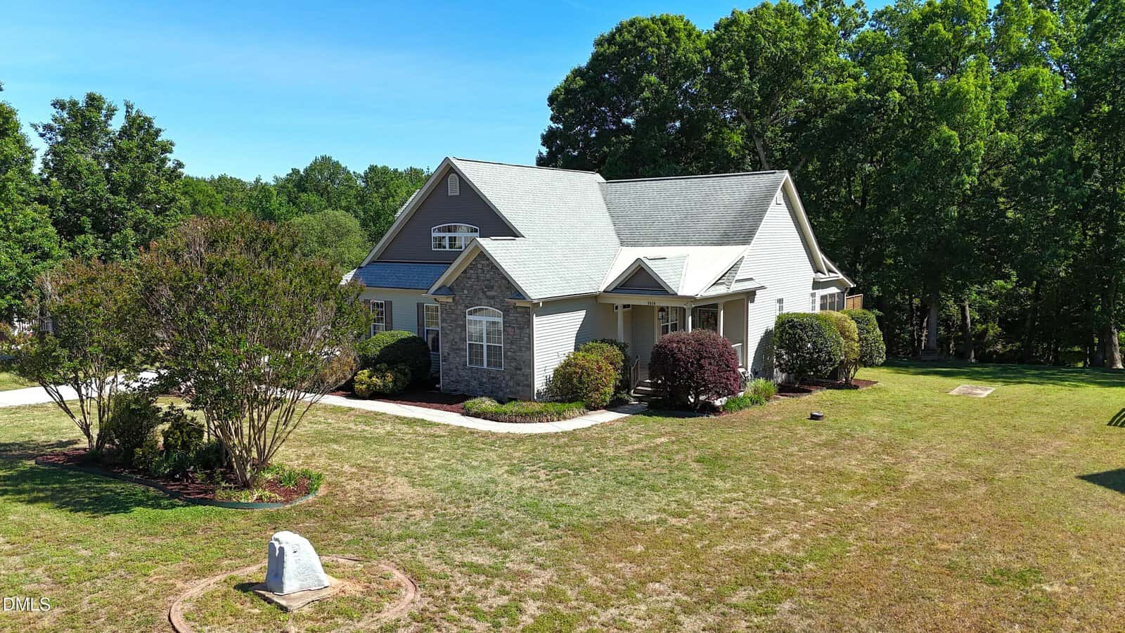 A light gray house with stone accents and a gabled roof sits on a grassy lawn with trees and landscaped bushes under a clear blue sky. A small concrete structure is in the front yard.