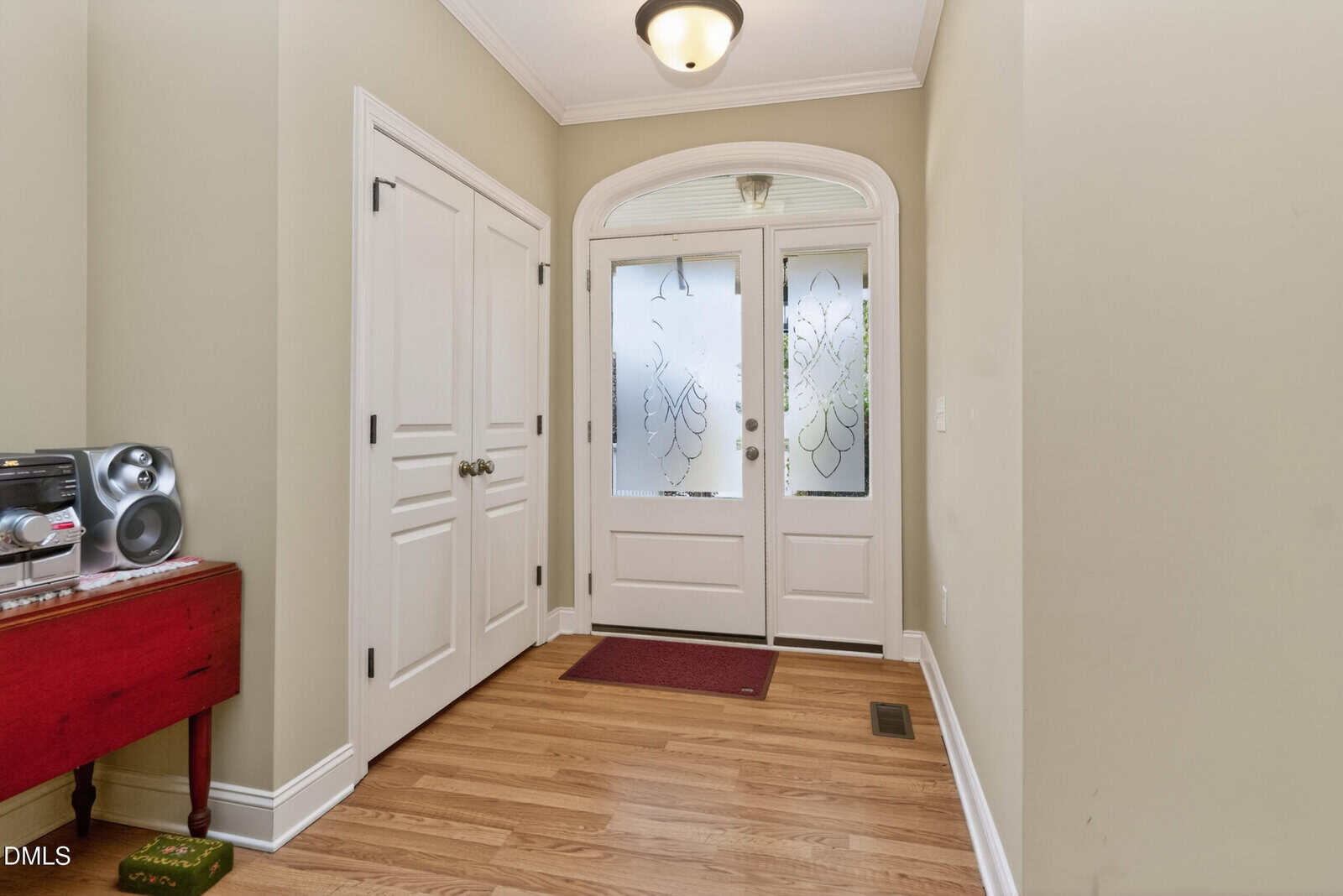 A hallway with light hardwood floors, double white doors on the left, a red table with a stereo, and a white front door with frosted glass panels featuring decorative patterns.