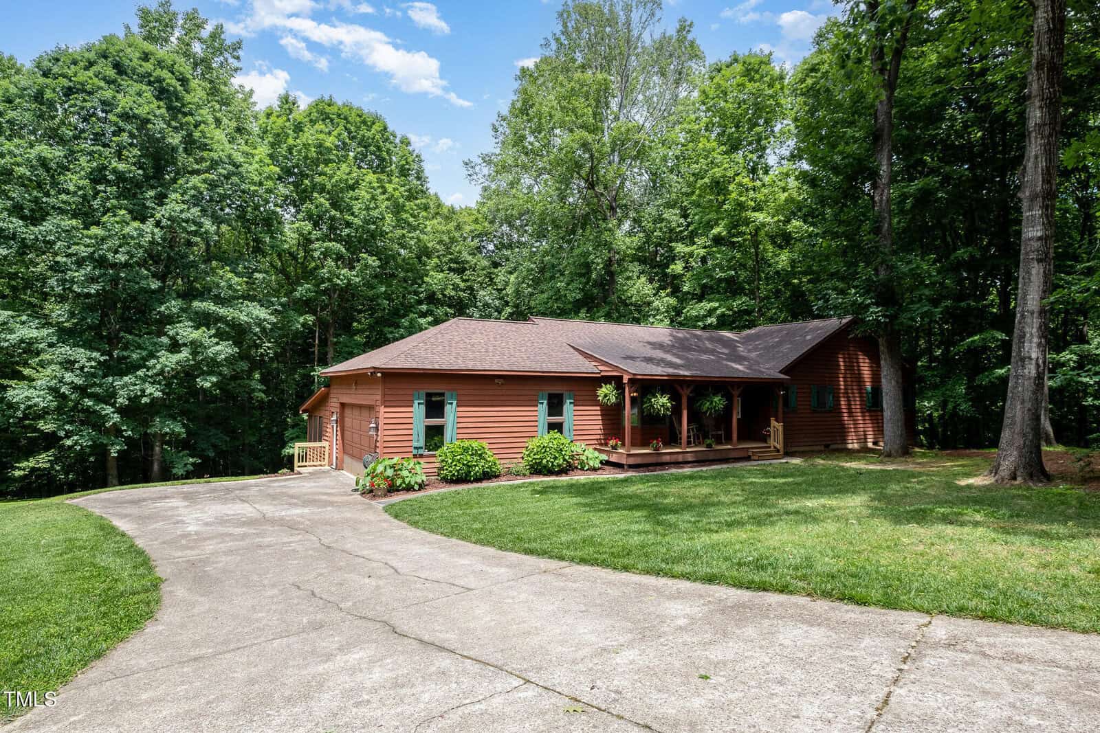A single-story log cabin with a covered front porch and hanging plants, surrounded by trees and greenery. A curved concrete driveway leads up to the house under a partly cloudy sky.