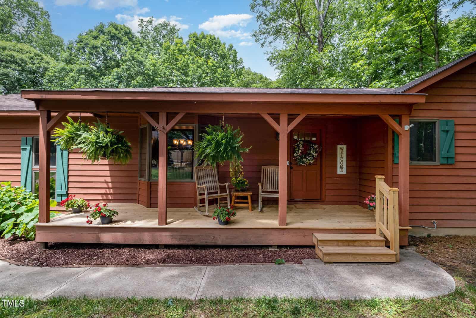 A cozy wooden house with a covered front porch, two rocking chairs, hanging ferns, potted flowers, and green shutters, surrounded by trees under a blue sky with clouds.