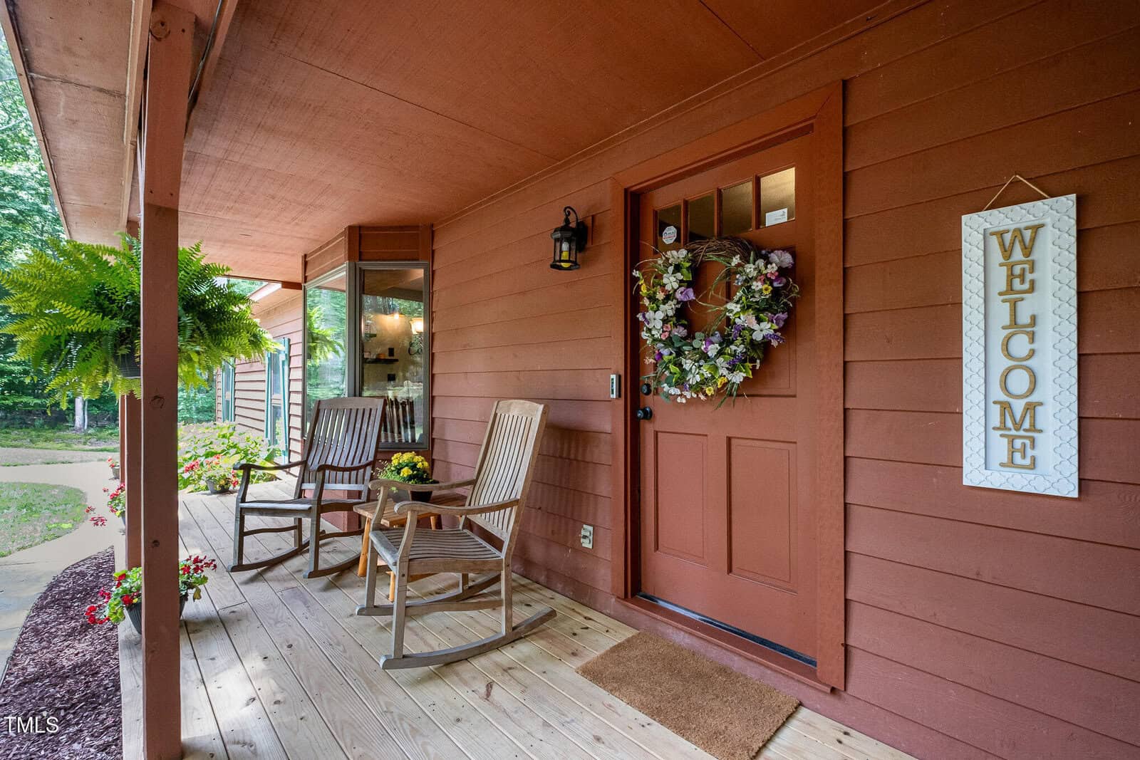 A cozy front porch with wooden flooring, two rocking chairs, potted plants, a welcome sign, and a front door decorated with a floral wreath. The porch is surrounded by greenery.