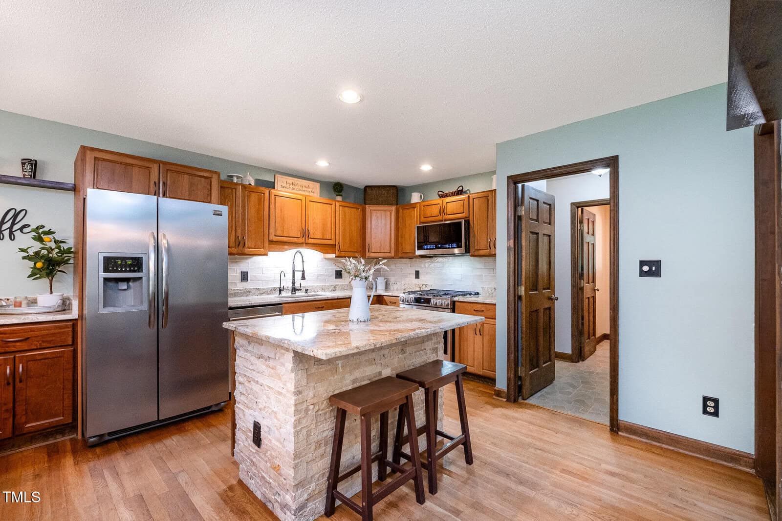Modern kitchen with stainless steel appliances, wooden cabinets, a central island with stone finish, two stools, and light wood flooring. Walls are light green and there is a doorway leading to another room.